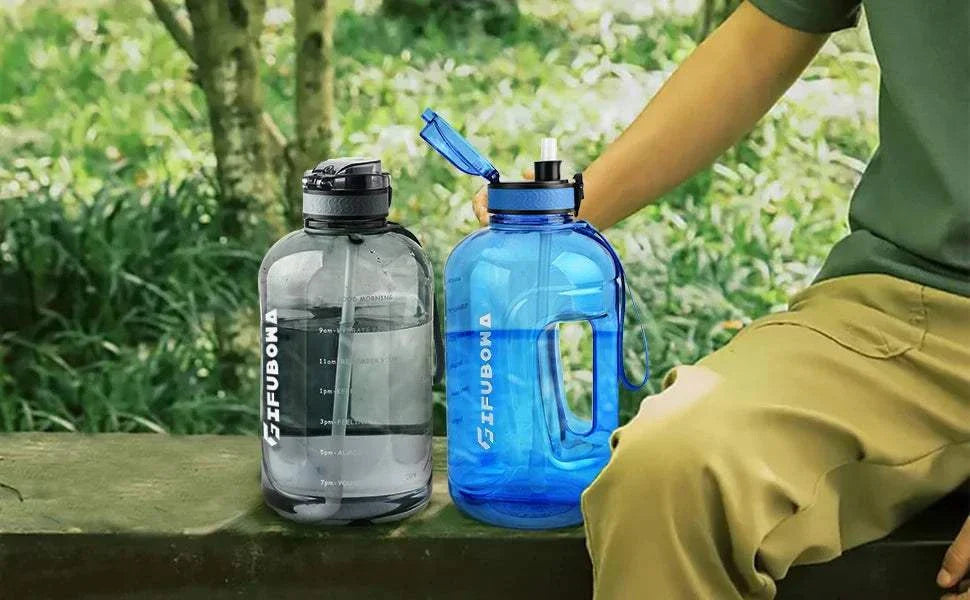 3L motivational water bottles with time markers, flip straw, and handle in blue and gray colors placed outdoors next to a person sitting on a bench.