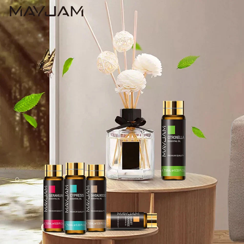 MAYJAM Essential Oil bottles with gold caps displayed with a reed diffuser on a wooden table surrounded by green leaves