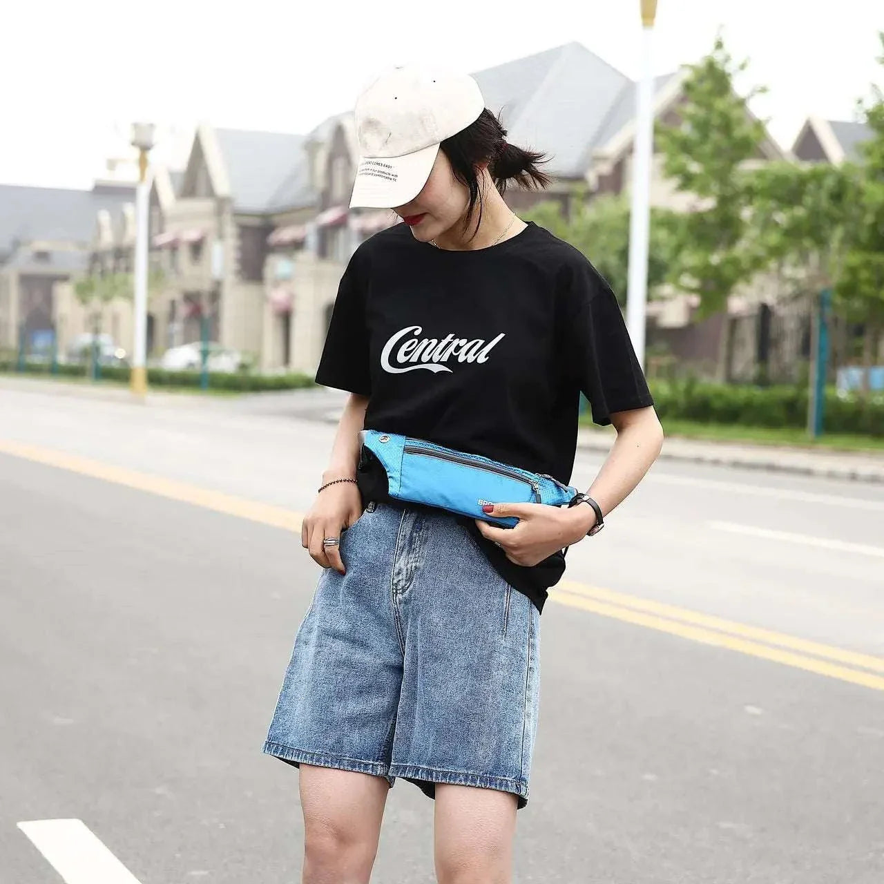 Woman wearing a blue nylon waist and chest bag over black t-shirt and denim shorts outdoors on street