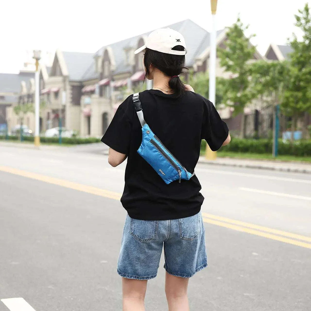 Person wearing a blue nylon waist and chest bag crossbody style with black shirt and denim shorts outdoors on a street with houses and trees background