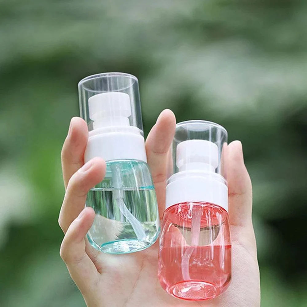 Hand holding two mini travel spray bottles with clear plastic caps, one filled with blue liquid and the other with pink liquid, outdoor background